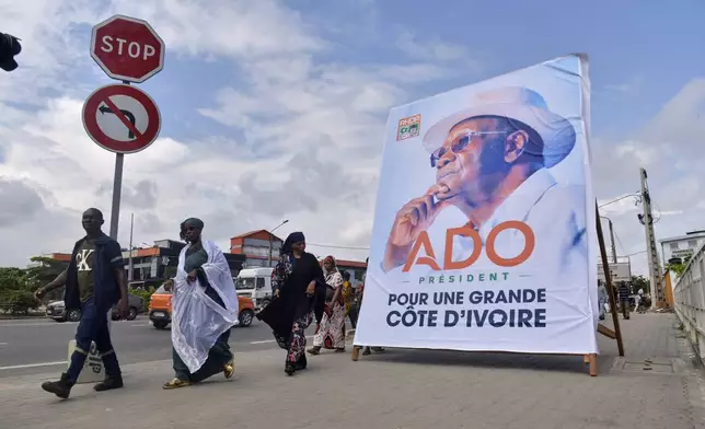 People walk past a campaign poster of President Alassane Ouattara in Abidjan, Ivory Coast, Friday, Oct. 10, 2025 (AP Photo/Diomande Ble Blonde)