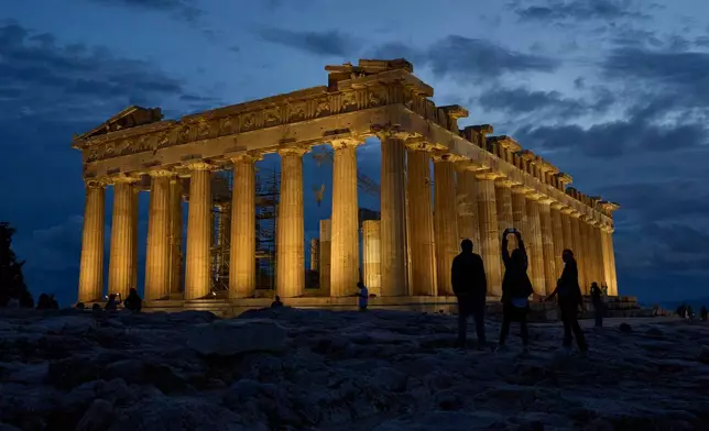 Tourists stand in front of the illuminated 5th century B.C. Parthenon temple atop the Acropolis hill in Athens, Friday, Oct. 31, 2025. (AP Photo/Petros Giannakouris)