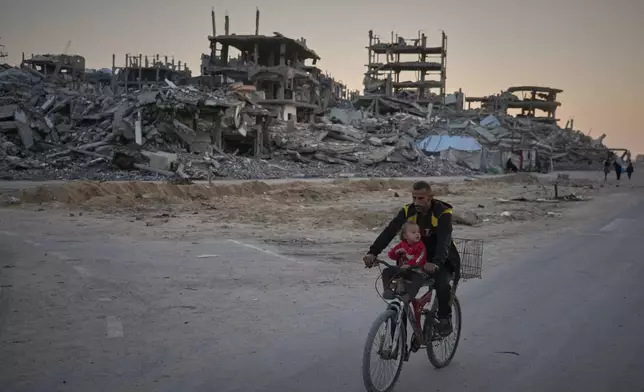 A man carries a child while riding his bicycle along a damaged street in the Al-Shati camp in Gaza City, Tuesday, Nov. 18, 2025. (AP Photo/Jehad Alshrafi)