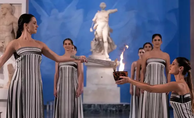 High Priestess Mary Mina, left, lights a torch from the Olympic flame during the flame lighting ceremony for the Milan Cortina 2026 Winter Olympics, at the archaeological museum of Olympia, Greece, Wednesday, Nov. 26, 2025. (AP Photo/Thanassis Stavrakis)
