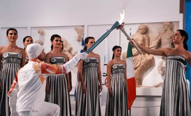 Actress Mary Mina, right, playing the role of high priestess, passes the Olympic flame to the first torchbearer Greek rower Petros Gaidatzis, during the flame lighting ceremony for the Milan Cortina 2026 Winter Olympics, at the archaeological museum of Olympia, Greece, Wednesday, Nov. 26, 2025. (AP Photo/Thanassis Stavrakis)