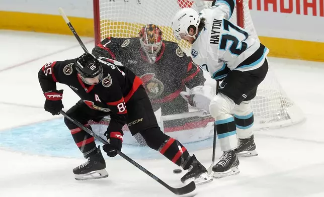 Ottawa Senators goaltender Leevi Merilainen (1) keeps his eye on the puck Utah Mammoth centre Barrett Hayton (27) stick handles the puck past defenceman Jake Sanderson (85) during first period NHL action, in Ottawa, Sunday, Nov. 9, 2025. (Adrian Wyld/The Canadian Press via AP)