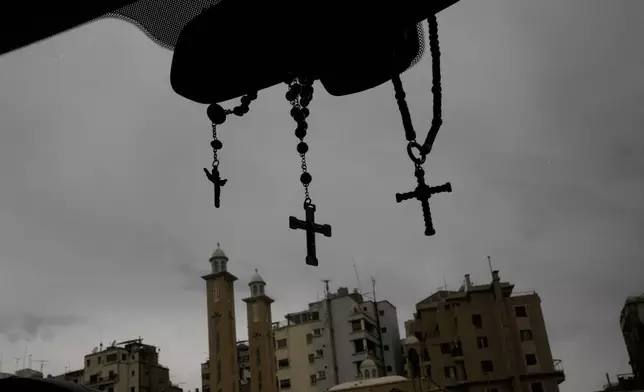 Rosaries and crosses hang from a car's rearview mirror as buildings and church towers rise in the background in Beirut, Lebanon, Friday, Nov. 14, 2025. (AP Photo/Hassan Ammar)