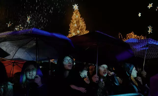 People listen to a concert during rainfall as Athens illuminated its Christmas tree at Syntagma Square on Thursday, Nov. 27, 2025. (AP Photo/Petros Giannakouris)