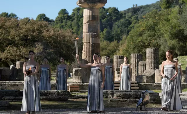 Actress Mary Mina, center, playing the role of high priestess holds the torch with the flame during a rehearsal ahead of the flame lighting for the Milan Cortina 2026 Winter Olympics, at the Ancient Olympia site, Greece, Monday, Nov. 24, 2025. (AP Photo/Thanassis Stavrakis)