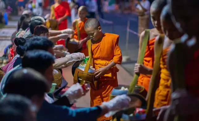 Novice Buddhist monks walk in a line seeking alms from residents and tourists waiting along a street before dawn in Luang Prabang, Laos, Friday, Nov. 7, 2025. (AP Photo/Eugene Hoshiko)
