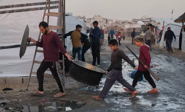 Displaced Palestinians walk through muddy pathways as a man and children carry cooking equipment at a camp in Jabalia, in the northern Gaza Strip, Sunday, Dec. 7, 2025. (AP Photo/Jehad Alshrafi)