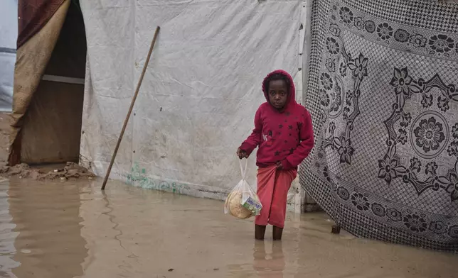 Carrying a plastic bag with bread, Tahreer Al-Rawagh, 8, looks on as he stands in a flooded area in a temporary tent camp after heavy rainfall in Deir al-Balah, central Gaza Strip, Friday, Dec. 12, 2025. (AP Photo/Abdel Kareem Hana)