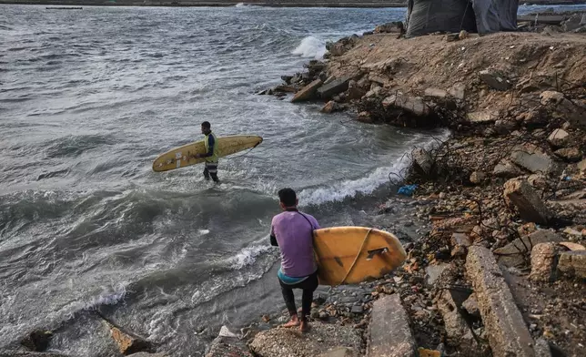 Palestinian Khalil Abu Jayyab, front, and Tahseen Abu Assi carry their boards as they enter the water along a damaged shoreline in Gaza City, Sunday, Dec. 28, 2025. (AP Photo/Jehad Alshrafi)