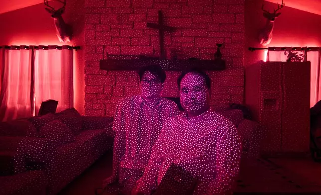Pastor Pan Yongguang, right, and his son Paul, members of a Chinese church living in exile after fleeing from China, are illuminated by cellphone infrared facial recognition beams while sitting for a photo in the community room of the ranch compound where they're living in Midland, Texas, Oct. 11, 2025. (AP Photo/David Goldman)