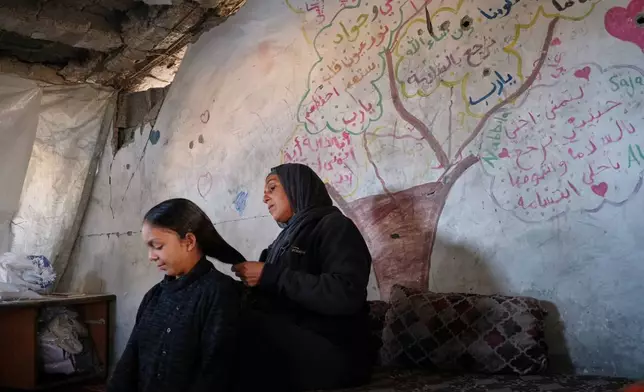 Amani Halawa combs her daughter Aya's hair under the roof of their damaged home, with a tree and messages of longing and hope painted behind them, in Jabaliya, Gaza City, Tuesday, Nov. 25, 2025. (AP Photo/Jehad Alshrafi)