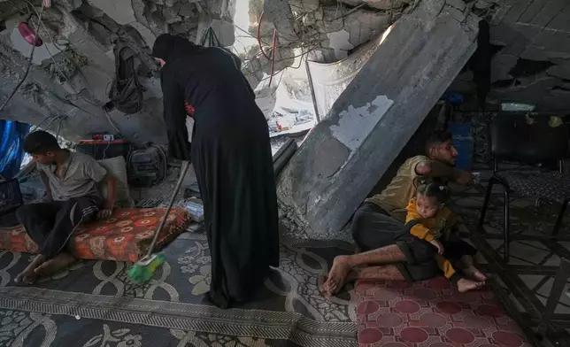 Sahar Taroush cleans her home beneath a partially collapsed roof while her children, Ammar, left, Hassan, right, and their 2-year-old sister, Ahlam, sit nearby, in Jabaliya, Gaza City, Tuesday, Nov. 11, 2025. (AP Photo/Jehad Alshrafi)