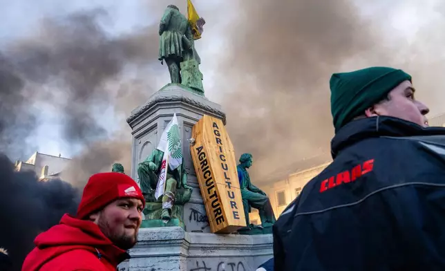 Two men walk by a wooden coffin placed on a statue base during a demonstration by European farmers outside the EU Summit meeting in Brussels, Thursday, Dec. 18, 2025. (AP Photo/Marius Burgelman)