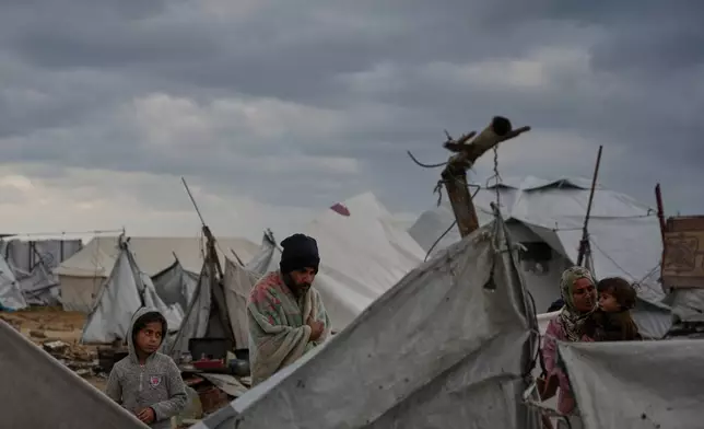 Wrapped in a blanket, Raafat Alwan, 32, stands outside his tent with his family in a makeshift camp for displaced Palestinians set up on the beach during a cold morning in Gaza City, Tuesday, Dec. 16, 2025. (AP Photo/Abdel Kareem Hana)