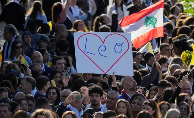 A woman raises a sign with a large red heart and the pope's name, "Leo," written inside as people wave Lebanese and Vatican flags while gathering to welcome Pope Leo XIV at the Psychiatric Hospital of the Cross in the town of Jal el-Dib, north of Beirut, Lebanon, Tuesday, Dec. 2, 2025. (AP Photo/Hassan Ammar)