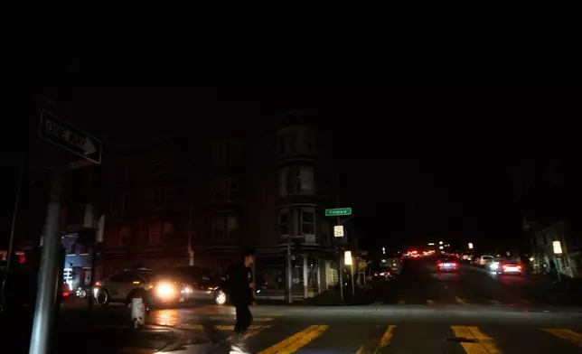 A pedestrian uses a flash light while crossing an intersection as power outages darken the streets, in San Francisco, Saturday, Dec. 20, 2025. (AP Photo/Jeff Chiu)