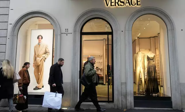 A man walks with a Prada shopping bag in front of a Versace shop, at the Montenapoleone luxury fashion street, in Milan, Italy, Tuesday, Dec. 2, 2025. (AP Photo/Luca Bruno)