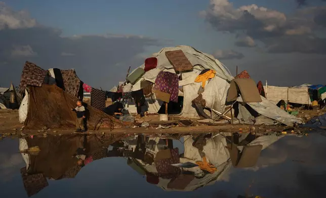 A Palestinian boy stands next to his mother washing clothes as mattresses hang to dry over their tent in a makeshift camp for displaced people set up on the beach in Gaza City, Tuesday, Dec. 16, 2025. (AP Photo/Abdel Kareem Hana)