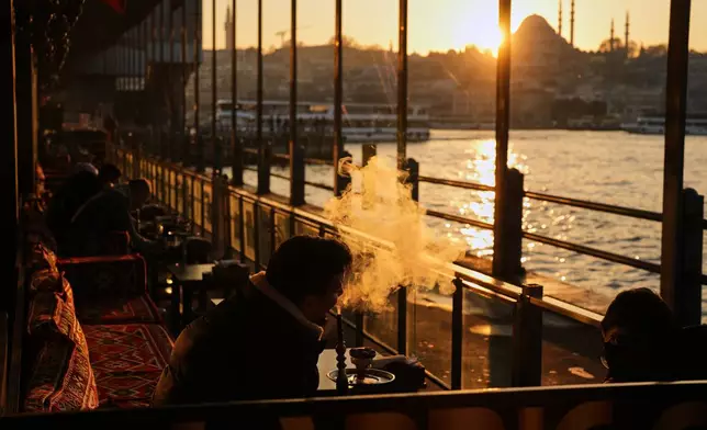 A man smokes a shisha water pipe at Galata bridge as the sun sets behind Suleymaniye mosque, in Istanbul, Turkey, Tuesday, Dec. 16, 2025. (AP Photo/Francisco Seco)