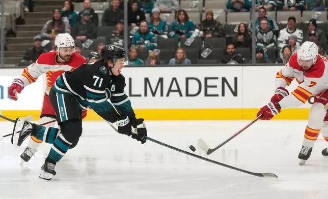 San Jose Sharks center Macklin Celebrini (71) reaches for the puck against Calgary Flames defenseman Rasmus Andersson, left, and defenseman Kevin Bahl (7) during the first period of an NHL hockey game in San Jose, Calif., Tuesday, Dec. 16, 2025. (AP Photo/Jeff Chiu)