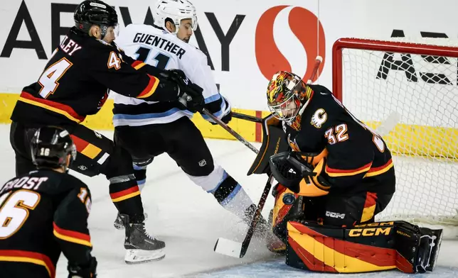 Utah Mammoth's Dylan Guenther, second from right, has his shot blocked by Calgary Flames goalie Dustin Wolf, right, as Flames' Joel Hanley, top left, checks him during third-period NHL hockey game action in Calgary, Alberta, Saturday, Dec. 6, 2025. (Jeff McIntosh/The Canadian Press via AP)