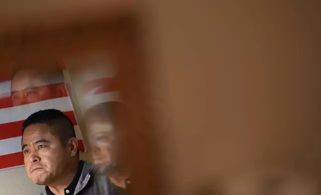 Former Chinese official Li Chuanliang is reflected in a mirror as he films a YouTube broadcast in his room alongside an American flag in the Mayflower Church community in Midland, Texas, Jan. 18, 2025. (AP Photo/Rebecca Blackwell)