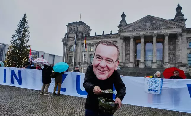 A demonstration with the mask of German Defence Minister Boris Pistorius attends a protest against the planned possibility of compulsory service in front of the German parliament in Berlin, Germany , Friday, Dec. 5, 2025. (AP Photo/Markus Schreiber)