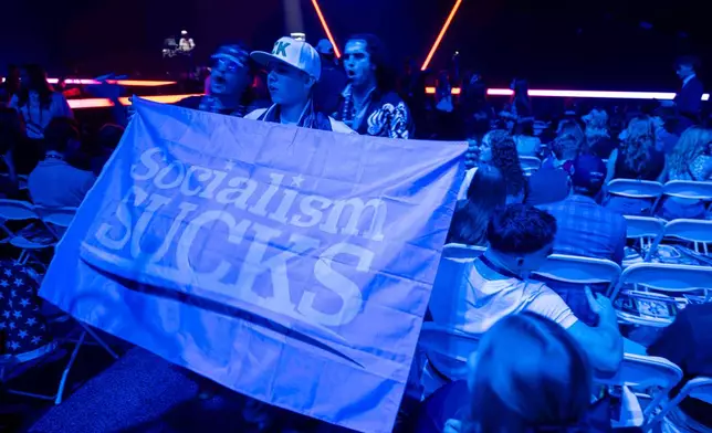 Students wave a flag reading "Socialism SUCKS" during Turning Point USA's AmericaFest 2025, Thursday, Dec. 18, 2025, in Phoenix. (AP Photo/Jon Cherry)