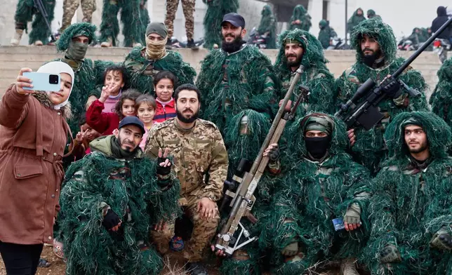 A child takes a selfie with soldiers of the new Syrian army dressed in camouflage as they gather before a parade, part of celebrations marking the first anniversary of the ousting of the Bashar Assad regime in Douma, on the outskirts of Damascus, Syria, Monday, Dec. 8, 2025. (AP Photo/Omar Sanadiki)