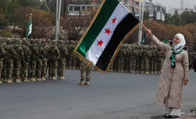 A woman waves the Syrian flag as members of the new Syrian army stand in formation during celebrations marking the first anniversary of the ousting of former President Bashar Assad in Damascus, Syria, Monday, Dec. 8, 2025. (AP Photo/Omar Sanadiki)