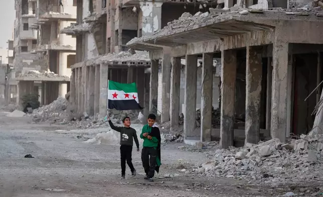 Two boys walk down a street carrying Syrian flags in the war-damaged Damascus suburb of Daraya, Syria, Friday, Dec. 5, 2025, ahead of celebrations marking the first anniversary of the ousting of the Bashar Assad regime. (AP Photo/Omar Sanadiki)