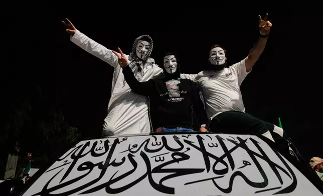 Syrian men wearing anonymous masks flash victory signs, as they stand on top of their car with its front window covered by an Islamic flag, during celebrations marking the first anniversary of the ousting of the Bashar Assad regime in Damascus, Sunday, Dec. 7, 2025. (AP Photo/Hussein Malla)