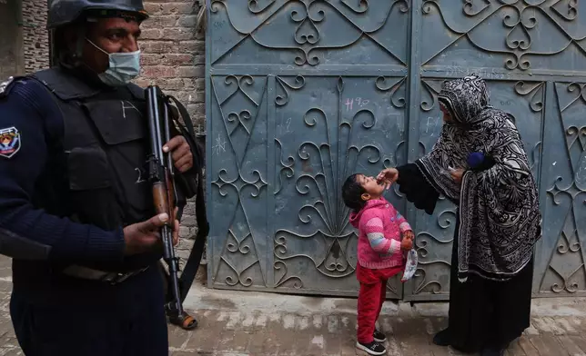 A police officer stands guard as a health worker, right, administers a polio vaccine to a child at a neighbourhood in Peshawar, Pakistan, Monday, Monday, Dec. 15, 2025. (AP Photo/Muhammad Sajjad)