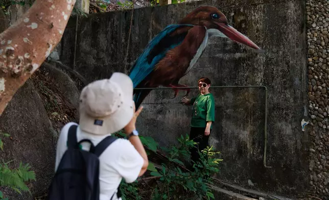 A visitor photographs her friend posing with a mural of a white-throated kingfisher painted on an abandoned house near Wang Tong village, Lantau, Hong Kong, Nov. 16, 2025. (AP Photo/May James)