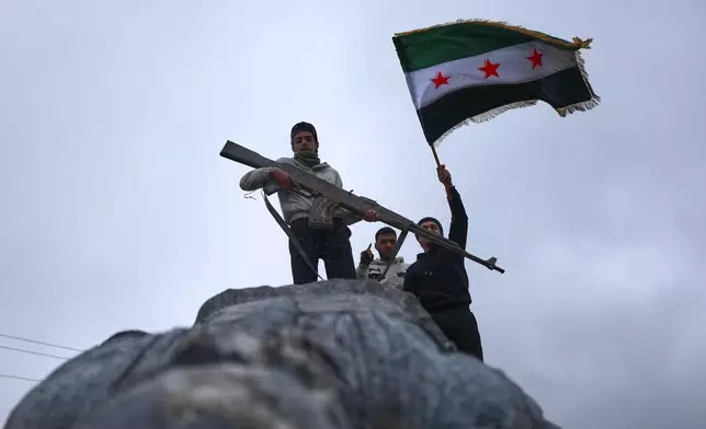 Residents wave a Syrian flag atop a toppled statue of a female Kurdish fighter, with one holding a statue's replica AK-47 that was part of the statue, after the takeover of the town by Syrian government forces from U.S.-backed Syrian Democratic Forces (SDF), in Tabqa, eastern Syria, Sunday, Jan. 18, 2026. (AP Photo/Ghaith Alsayed)
