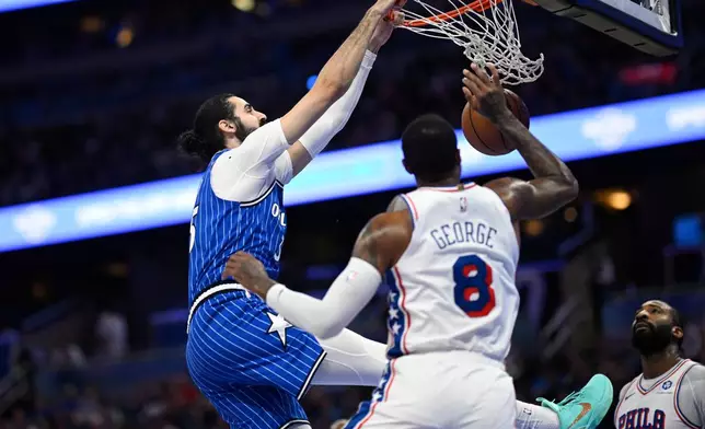 Orlando Magic center Goga Bitadze, left, dunks between Philadelphia 76ers forward Paul George (8) and center Andre Drummond, right, during the first half of an NBA basketball game, Friday, Jan. 9, 2026, in Orlando, Fla. (AP Photo/Phelan M. Ebenhack)