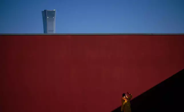 A woman visiting a recreation park at a rooftop of a shopping mall poses against a red wall as skyscraper China Zun Tower is seen behind in Beijing, China, Tuesday, Jan. 20, 2026. (AP Photo/Andy Wong)