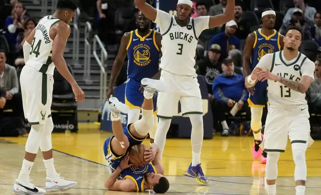 Golden State Warriors guard Stephen Curry, bottom, rolls on his back while holding onto the ball between Milwaukee Bucks forward Giannis Antetokounmpo, left, center Myles Turner (3) and guard Ryan Rollins (13) during the second half of an NBA basketball game in San Francisco, Wednesday, Jan. 7, 2026. (AP Photo/Jeff Chiu)