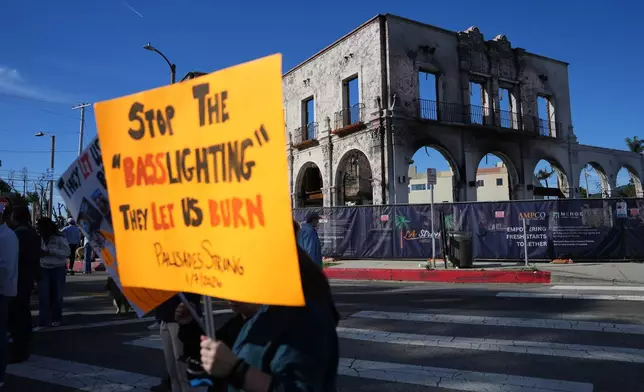 Demonstrators hold signs in front of a fire-ravaged building on the one-year anniversary of the Palisades Fire in the Pacific Palisades neighborhood of Los Angeles Wednesday, Jan. 7, 2026. (AP Photo/Jae C. Hong)
