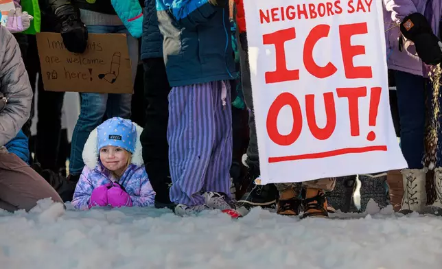 A child sits in the snow as demonstrators hold signs during a news conference at Lake Hiawatha Park in Minneapolis, on Friday, Jan. 9, 2026, demanding Immigration and Customs Enforcement be kept out of schools and Minnesota following the killing of 37-year-old mother Renee Good by federal agents earlier on Wednesday. (Kerem Yücel/Minnesota Public Radio via AP)