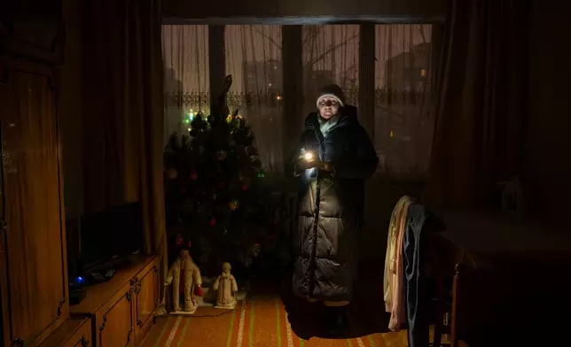 Lyudmila Kopal wearing warm clothes shines a flashlight as she stands by a Christmas tree during a blackout caused by Russia's regular air attacks against the country's energy in Kyiv, Ukraine, Tuesday, Jan. 13, 2026. (AP Photo/Danyil Bashakov)