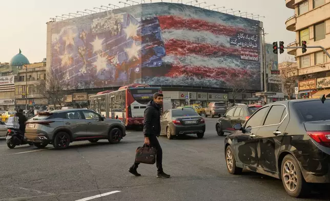 A man walks among vehicles in front of a billboard depicting a damaged U.S. aircraft carrier with disabled fighter jets on its deck and a sign reading in Farsi and English, "If you sow the wind, you'll reap the whirlwind," at Enqelab-e-Eslami (Islamic Revolution) Square in Tehran, Iran, Sunday, Jan. 25, 2026. (AP Photo/Vahid Salemi)