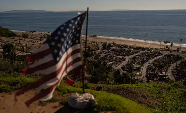 A tattered U.S. flag flaps in the wind over the remains of a mobile home park that was destroyed in the Palisades Fire along the Pacific Ocean, Friday, Dec. 5, 2025, in the Pacific Palisades neighborhood of Los Angeles. (AP Photo/Jae C. Hong)
