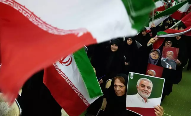 Women wave national flags as they hold up posters of the late commander of the Iran's Revolutionary Guard expeditionary Quds Force, Gen. Qassem Soleimani, who was killed in a U.S. drone attack in 2020 in Iraq, during a ceremony commemorating his death anniversary at the Imam Khomeini grand mosque in Tehran, Iran, Thursday, Jan. 1, 2026. (AP Photo/Vahid Salemi)
