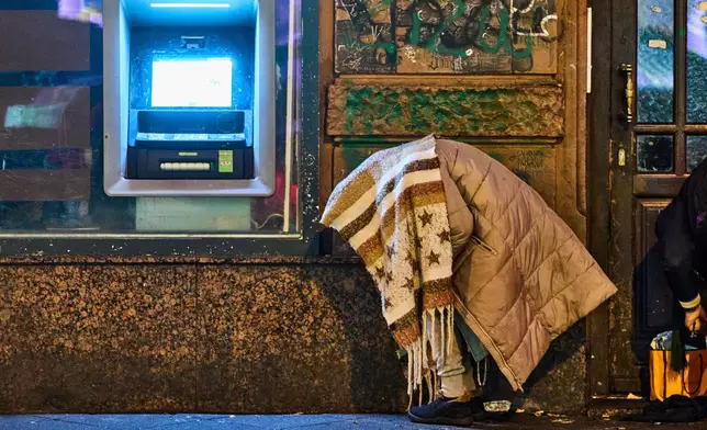 A woman covers herself with a blanket against the cold next to a banking machine in Frankfurt, Germany, Wednesday, Jan. 7, 2026. (AP Photo/Michael Probst)