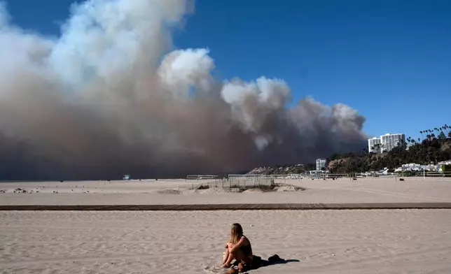 FILE - A lone sunbather sits and watches a large plume of smoke from a wildfire rise over the Pacific Palisades, in Santa Monica, Calif., Jan. 7, 2025. (AP Photo/Richard Vogel, File)