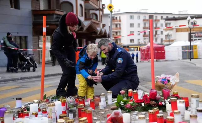 A police officer helps a boy to light a candle near the sealed off Le Constellation bar in Crans-Montana, Swiss Alps, Switzerland, Friday, Jan. 2, 2026, where a devastating fire left dead and injured during the New Year's celebrations. (AP Photo/Baz Ratner)