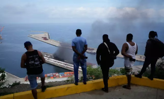 Men watch smoke rising from a dock after explosions were heard at La Guaira port, Venezuela, Saturday, Jan. 3, 2026. (AP Photo/Matias Delacroix)