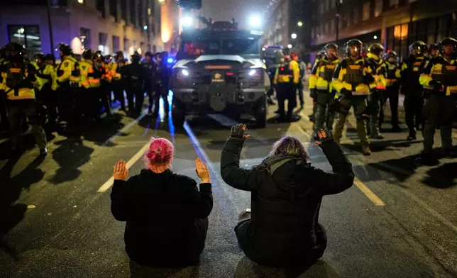 Two people sit in the street with their hands up in front of Minnesota State Patrol during a protest and noise demonstration calling for an end to federal immigration enforcement operations in the city, Friday, Jan. 9, 2026, in Minneapolis. (AP Photo/John Locher)
