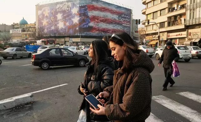 Two girls, not wearing the legally required headscarves, walk past a billboard depicting a damaged U.S. aircraft carrier with disabled fighter jets on its deck and a sign reading in Farsi and English, "If you sow the wind, you'll reap the whirlwind," at Enqelab-e-Eslami (Islamic Revolution) Square in Tehran, Iran, Sunday, Jan. 25, 2026. (AP Photo/Vahid Salemi)